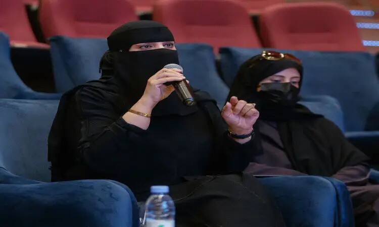 سيدة ترتدي نقابًا أسود وتتحدث في ميكروفون أثناء جلوسها على الأزرق في جامعة القصيم، بالإضافة إلى جانبها تجلس امرأة أخرى مرتدية كمامة وكمامة. للحفاظ على حصيرة الفراغات وزجاجة الماء في المكان، مما يدل على فعالية "التحول في التعليم" المقامة احتفاءً بـ "اليوم العالمي للمعلم". كما تميز الحضور بارتداء الشماغ السعودي والعقال والبشت السعودي، بما في ذلك يبرز الطابعة الرسمية والهوية الوطنية للفعالية.