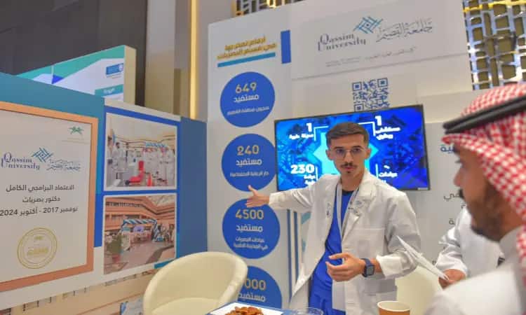 رجل يرتدي غترة سعودية وعقالاً وبشتاً سعودياً إلى جانب شماع أبيض (معطف مختبر) وهو يشرح للحضور بجناح جامعة القصيم خلال المؤتمر السعودي للبصريات، تمييز لشاشة تعرض إحصائيات ومعلومات باللغتين العربية والإنجليزية في مكان داخلي مضاء بشكل ساطع.