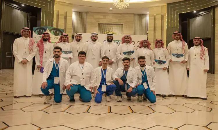 مجموعة من الرجال المؤثرين باللون الأسود فوق البشت السعودي، بالإضافة إلى الشماغ والعقال، يصطفون سوياً في صورة داخلية جماعية أثناء فعاليات المؤتمر السعودي للبصريات في القصيم. يؤمن في الخلفية ملصقات تحمل طابعاً بصرياً ورسومات كبيرة للعين، مما يدل على دقة التوقيع والموافقة بالمظهر الوطني الرسمي.