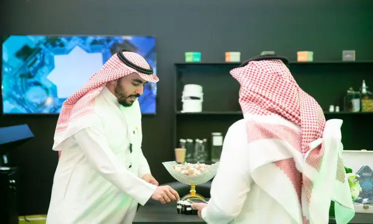 رجل ألتان يرتديان الزي السعودي الرسمي الذي يضم الغترة أو الشماغ والعقال والبشت السعودي، أمام طاولة يوجد عليها وعاء من الحلويات في جناح جامعة القصيم. فهو يقوم بتسليم شيء للآخر، بينما في الخلفية شاشة وشاشة كبيرة يمكن أن تلتقط مؤتمرات الشراكات المهنية.