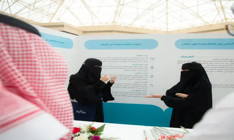 تتواصل سيدتان خارجان أعضاء كاملة في جناح المعلومات الخاصة بعمادة شؤون الطلاب في جامعة القصيم، حيث يبرز برنامج رواد. الرجل بجانبهما يرتدي البشت السعودي، والشماغ والعقال. يوجين مازينًا بعبارات بالعربية وزهور، دعمًا للأشخاص ذوي الشهرة العالمية.