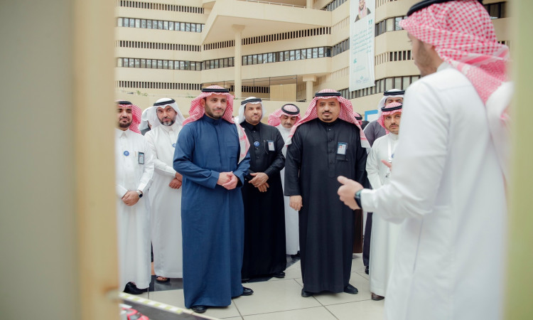 مجموعة من الرجال الحاضرين الغترة والشماع والعقال والبشت السعودي يجتمعون في معمل كود جامعة في القصيم، ويستمعون إلى المتحدث الرسمي. وعكس شرفات المبنى الحديث للطابعة العصرية المناسبة، والتي تشمل ريادة الأعمال الرقمية الشاملة.