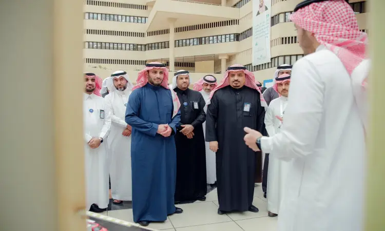 مجموعة من الرجال الحاضرين الغترة والشماع والعقال والبشت السعودي يجتمعون في معمل كود جامعة في القصيم، ويستمعون إلى المتحدث الرسمي. وعكس شرفات المبنى الحديث للطابعة العصرية المناسبة، والتي تشمل ريادة الأعمال الرقمية الشاملة.