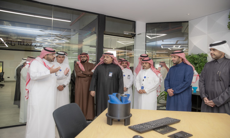 مجموعة من الرجال مرتدين الغترة السعودية أو الشماغ والعقال والبشتاء السعودي، إلى جانب الضيوف بدلت، في أحد البنات بمعمل كود الجامعة في جامعة القصيم. يستمتع الجميع باهتمام لا أحد يحيط بهم من جدران ومكاتب عمل ونباتات خضراء، بما في ذلك يعكس واقع عمل تتطلعات ريادة الأعمال الرقمية.