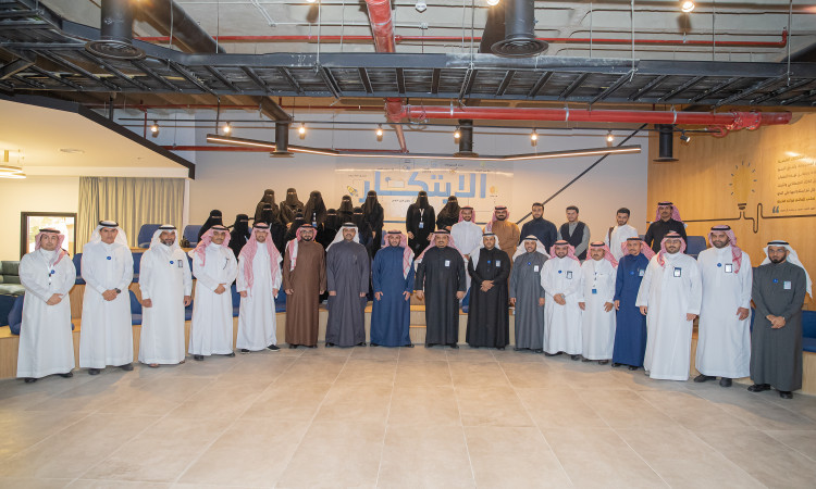 مزيج كبير من الرجال والنساء تألق الغترة أو الشماغ والبشت السعودي في صورة جماعية للسيدات بمعمل كود الجامعة، حاضر في الخلفية النصية عربي على الجدار بوضوح التزام هيئة الاتصالات والفضاء والتقنية بريادة الأعمال الرقمية في بيئة حديثة ضمن جامعة القصيم.