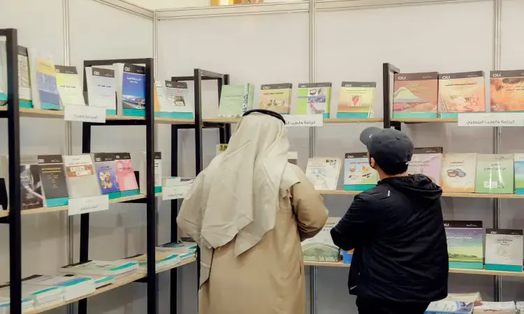 للاتصالان يرتديان الغترة والعقال والبشتاء السعودي امام رفوف معدنية سوداء تزينها اصدار الجامعة، وسط معرض القصيم للكتاب. تتراص الكتب والمجلات العربية على الرفوف المقامة بجدار أبيض، ما يعكس علميا ومعرفية متميزة تليق بمكانة جامعة القصيم.