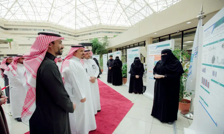 تجمع عدد من الرجال النساء مرتدين الغترة أو الشماغ مع العقال، بالإضافة إلى البشت السعودي، متابعة إحدى الباحثينات المرقابية أثناء تقديمها لملصق بحثي ضمن فعاليات ملتقى طلبة الدراسات العليا في القصيم، وذلك في قاعة داخلية تحت قبة مميزة التصميم.