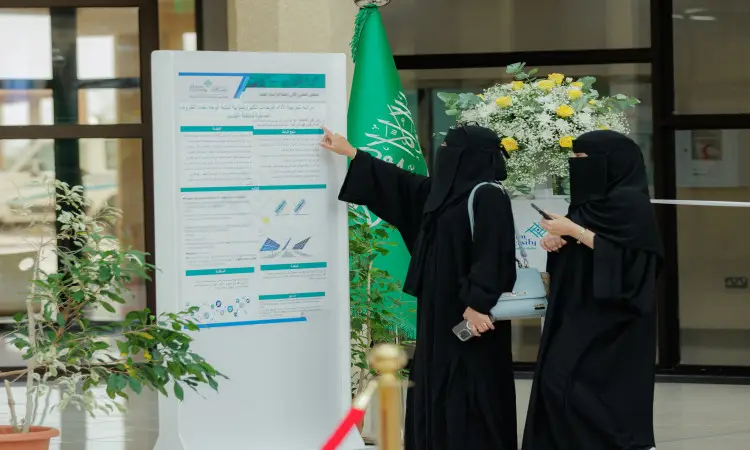 أمرأتان ترتديان رداءات بيضاء وبشت سعودي مع غترة وعقال، فلوريداان أمام لوحة معلوماتية عن ملتقى قمة الدراسات العليا في جامعة القصيم، ويانان إلى محتوى اللوحة بينما تحمل إحداهما هاتفًا. يظهر في الخلفية علم المملكة العربية السعودية وتنسيق ورد ونباتات داخلية، مما يضفي على المكان مسؤولاً تأويل هوية الجامعة.