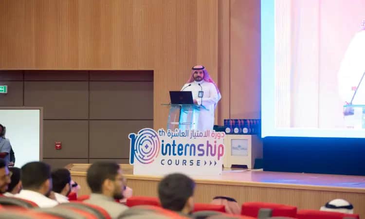 رجل يفضل البشتاء السعودي والغترة والعقال، بالحديث من على منصة الخطابة خلال فعاليات الدورة العاشرة للامتياز لطلاب الطب، وذلك كما يظهر في الشاشة الكبيرة المجاورة له. ويجلس في المقدمة عدد من الحضور منسوبي جامعة القصيم.