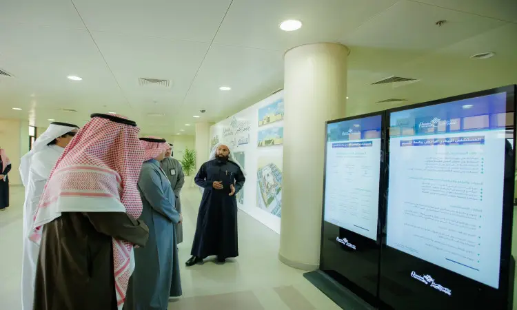 يجتمع مجموعة من الرجال مرتدين الغترة أو الشماغ والعقال والبشت السعودي، بينهم معالي وزير التعليم، للاستماع إلى مقدمي يرتدون بشتاً داكناً، وذلك خلال متابعتهم لشاشات رقمية كبيرة تعرض نصوصاً عربية، في بيئة داخلية حديثة ضمن أروقة جامعة القصيم.