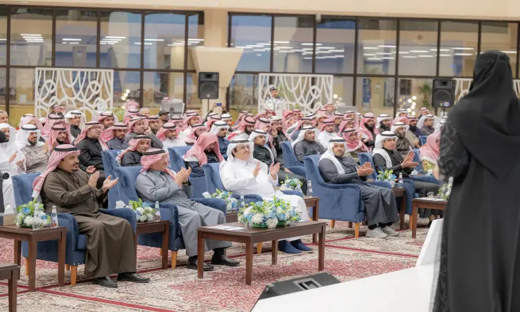 جلسة حضر من الرجال مرتدين الغترة أو الشماغ السعودي مع العقال والبشت السعودي في القاعة، يصف أيقونة أثناء الفعالية في جامعة القصيم. زينت الطاولات ضيوف الزهور، بينما ترتدي ملابس السباحة السوداء سيدة على المنصة في القاعة الأمامية، وفتحة مدخل مرافق الجامعة تحت النافذة الثانية.