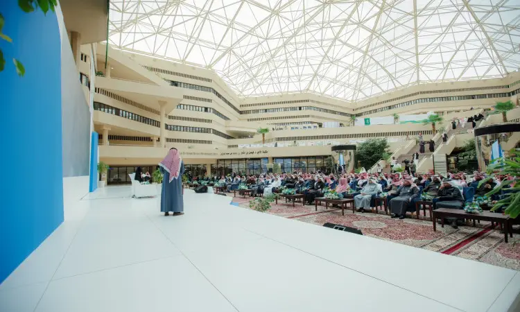 رجل يرتدي الزي السعودي التقليدي، متشحًا بغتة سعودية بيضاء وقال وبشت سعودي، يُحتمل أنه معالي وزير التعليم، يلقي كلمة على منصة أمام جمهور كبير يجلس في قاعة مرحبا بكة عند القصيم، إلا بسقف زجاجي تصميمي وعدة شرفات.