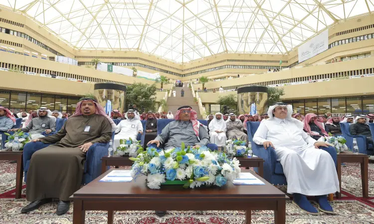 يجلسون مجموعة من الرجال ينتظرون الغترة أو الشماغ السعودي مع العقال والبشت السعودي، ومن بينهم معالي وزير التعليم، في الصف الأول خلال المخيم الكبير أُقيمت في رحاب جامعة القصيم، حيث يحيط بهم جمع غفير من الحضور فيما تزيّن مرافقة القاعة تنسيقات الزهور البيضاء والزرقاء.