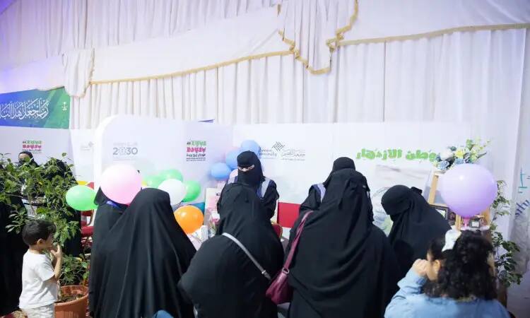 A group of women wearing black abayas and niqabs at one of the Buraydah Spring Festival stands on campus, which was decorated with balloons and natural plants. A small child appeared near the platform, with Arabic-language signs for students belonging to a club in the background. Some attendees participated in the event in the presence of the Saudi Shemagh or Ghutrah with the Saudi Aqal and Bisht, which added to the event of an official, recognizing the prestige of Qassim University and its keenness to preserve national values and traditions.