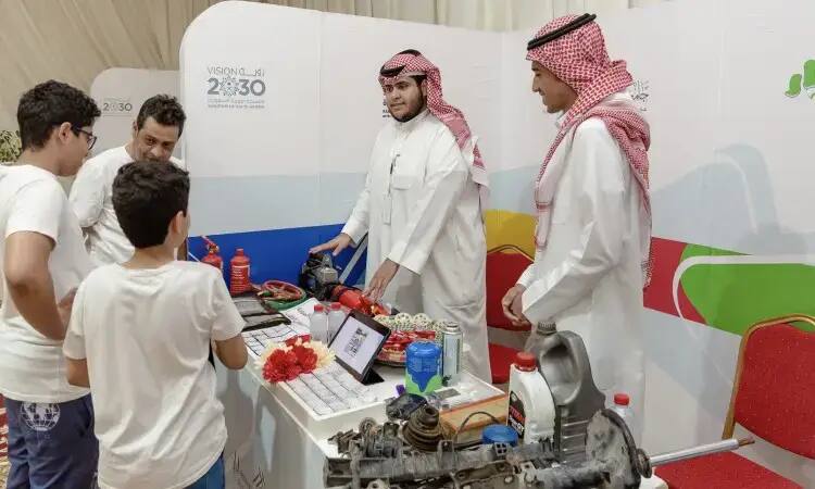 Two men dressed in Saudi Arabia's Ghutrah, Aqal and Bisht behind a table displaying smart game mechanics, chatting with three students in front of an awareness corner that squeezed the Vision 2030 slogan. This activity is part of the activities of the Qassim University Student Club participating in the Buraydah Spring Festival.