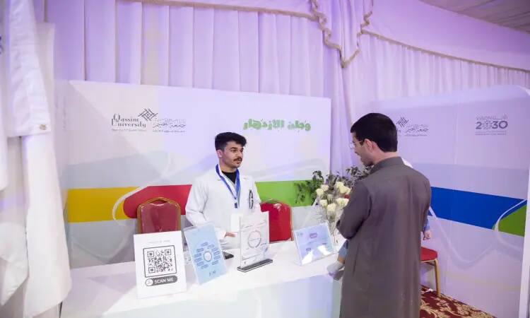A man wearing a white coat behind the wings of the Buraydah Spring Festival logo and Qassim University, talking to another man wearing a Saudi ghutra and a Saudi besht as part of the men's event. The flight displays models of Nasdin and the Saudi Vision 2030 logo, as well as a QR code (Quick Response Code).