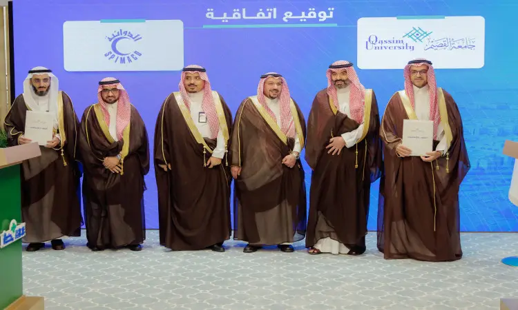Six men, resplendent in shemagh or ghutra and aqqal, and wearing the Saudi bisht, stand at the women's podium, holding the documents of cooperation and smiling. Behind them is a blue background with logos and texts in Arabic, as well as the name of Qassim University, during the signing ceremony of cooperation agreements under the patronage of His Highness the Emir of Qassim.
