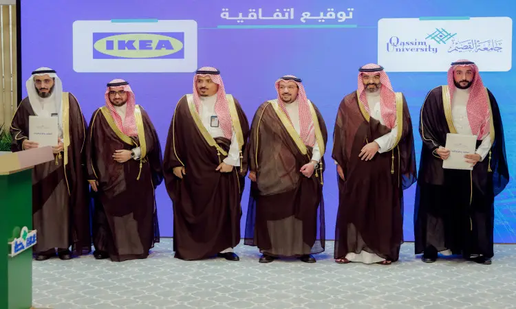 A group of six men in Saudi Arabian ghutra or shemagh with Saudi Arabian headband and bisht gather in front of a screen displaying the logos of Qassim University and IKEA, holding documents during the signing ceremony to formalize cooperation.