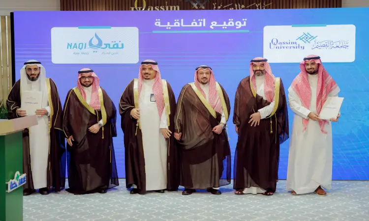 Six men, resplendent in the Saudi ghitra or shemagh and aqal, complete with Saudi bisht, in front of a screen displaying "Qassim University" and "Pure Water Company." The men sign a document signaling cooperation. On the screen is a text in Arabic that reads "Emir of Qassim Region."