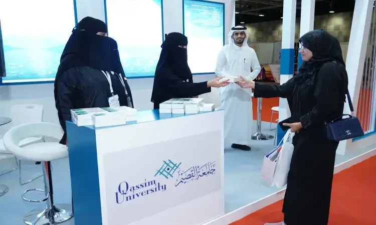ثلاث سيدات ترتدين العباءة والنقاب تتن خلف جناح جامعة القصيم في المعرض العالمي للتعليم العالي، ويسلمن مطوية لترتدي سترة. الرجل في الخلف يرتدي الشماغ السعودي والعقال والبشت السعودي مبتسمًا.
