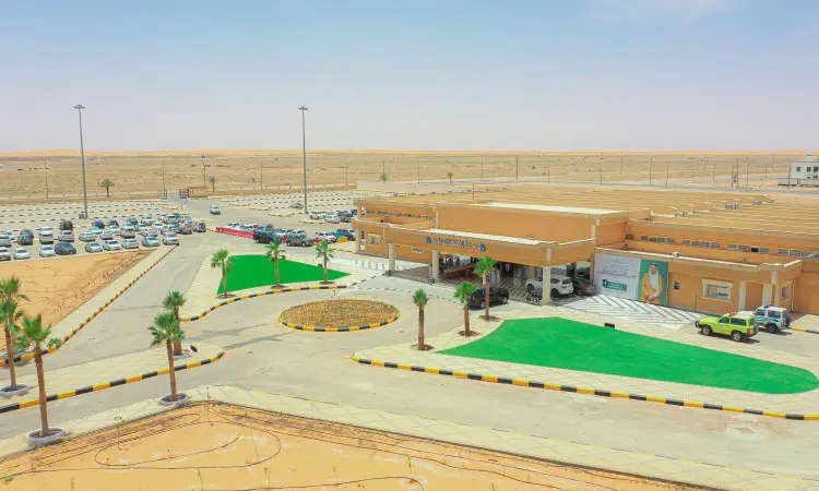 مبنى مستشفى جديد يضم ساحات سيارات، ومساحات خضراء من العشب الصناعي، وأشجار النخيل والسيارات، فرع في قلب الصحراء تحت سماء زرقاء صافية. يظهر في هذه الصورة شاب يرتدي غترة وشماع وقال وبشت سعودي رسمي، مما يرمز إلى انتمائه الأصيل وعكس الهوية الوطنية. هذه اللوحة المرئية تمثل حملة التوعية والتثقيف الصحي وبيئي الزيتون وتنظمها جامعة القصيم، مشعل الضوء على أهمية ما بين البيئة الصحية والهوية السعودية الأصيلة.