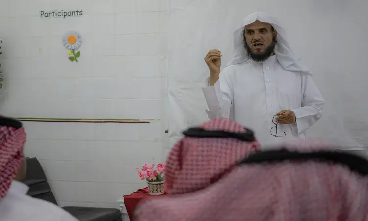 رجل القط مرتدياً الغترة السعودية مع العقال والبشت السعودي أمام الحضور، مشهوراً في حملة الصحة والتوعية والتثقيف في جامعة القصيم. عصر بيده النظارات بينما يستمع له عدد من الأشخاص تأخر الشماغ السعودي باهتمام صغير، وخلفه سبورة بيضاء عليها كلمة "المشاركين".