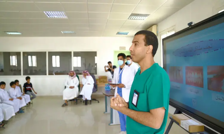 يقوم رجل بتقديم زي طبيًا أخضر معلومات توعوية عن صحة الفم والأسنان عبر شاشة عرض جماعية لعدد من الطلاب وعدد من أولياء الأمور الجالسين في القاعة. وتؤكد بالمشهد أعضاء من المحاربين العسكريين الماردين المؤثرين البيض والكمامات، بالإضافة إلى عدد من الحضور بالزي السعودي الرسمي، متوشحين الكتيرة أو الشماغ والعقال، وبعضهم مرتدياً البشت السعودي بما في ذلك تأكيد كامل على أنها جامعة القصيم.