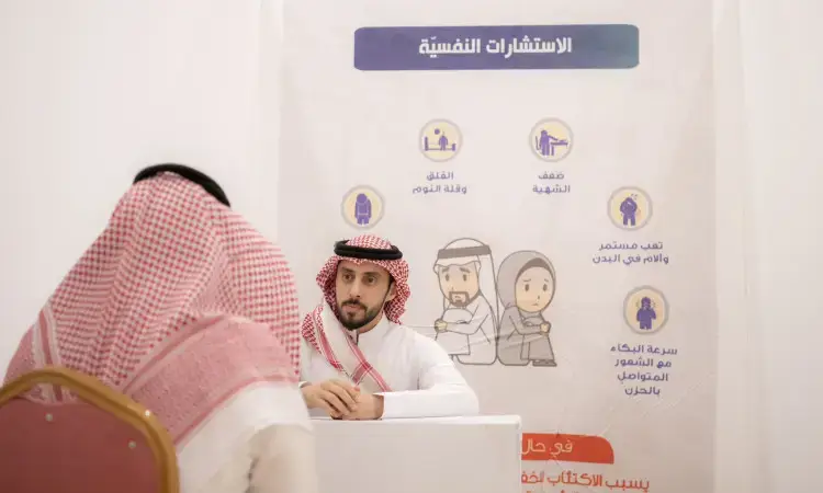 The man wearing the Saudi Ghutrah, Eqal and Bisht sits at the table, talking to another man whose back is to the camera. In the background is a poster that highlights the importance of the psychological campaign, focusing on health awareness and education among the staff and students of Qassim University.