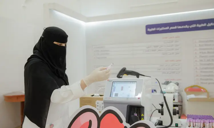 Dressed in a niqab with a headband, as well as a formal Saudi besht, a Saudi woman endures a tube test while working with lab equipment in a clinical environment at Qassim University to promote health awareness. Behind her on the wall is a chart with Arabic text.