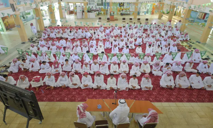 A large number of young men, dressed in the Saudi ghutra or shemagh and aqal, as well as the traditional Saudi bisht, gather in a red-carpeted mosaic hall. They sit respectfully and listen to three frontline players. Hanging above them are green banners with messages of awareness and education. Shoes are laid out in an organized carpet pattern at the edges, as befits the formal atmosphere of Qassim University.