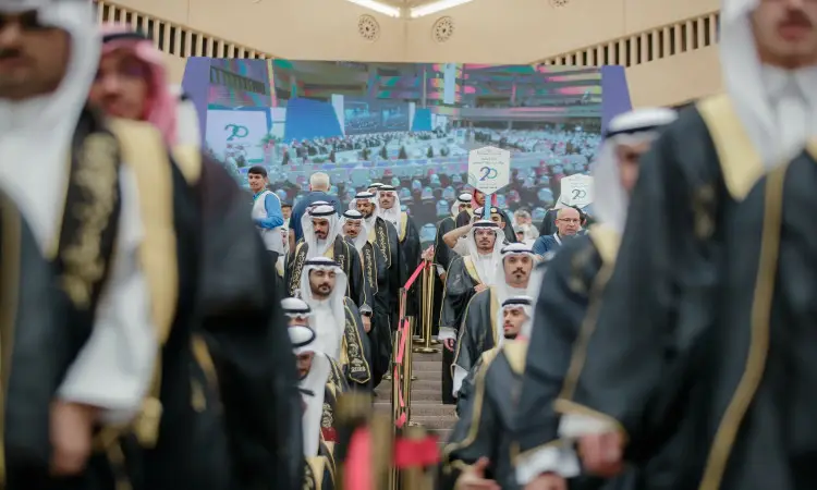 مجموعة من الرجال الحاضرين الغترة السعودية أو الشماغ والعقال والبشت السعودي، يهبطون الدرج خلال خريج الجامعة. فترة التوقف لافتات تعبيرية. المحتسب في شاشة العرض الكبيرة صورة لجمهور يحتفي بتخرج الطلاب، وذلك تحت رعاية كريمة سمو الأمير الدكتور فيصل بن مشعل بن سعود بن عبدالعزيز أمير منطقة القصيم.