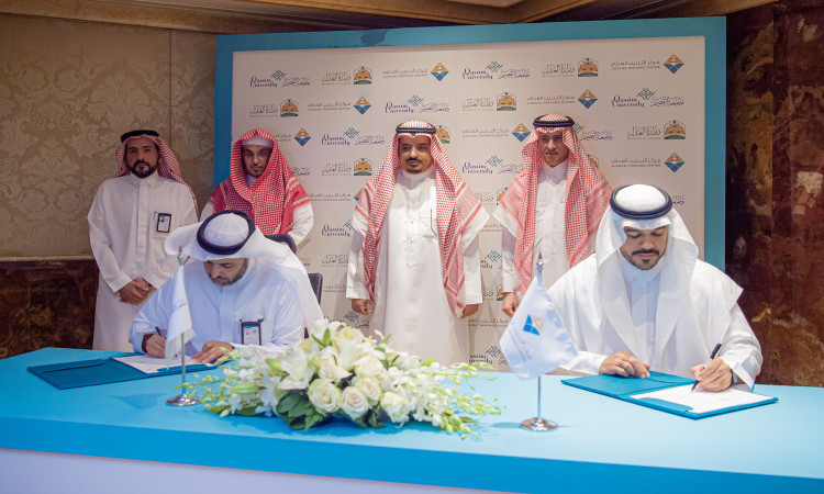 يوقعتان على مستندات فوق مستندات طاولة الزينة بالزهور البيضاء والأعلام الصغيرة، بينما الاتصال خلفهما أربعة رجال الغترة أو الشماغ والعقال، ويكتمل ظهور العنصر بالبشت الرسمي السعودي. وتبرز في الخلفية شاشة تحمل شعار التعاون بين مركز التدريب العدلي ووزارة العدل، وسط احترام السيدات الحرص على جامعة القصيم على تعزيز الشراكات الوطنية.