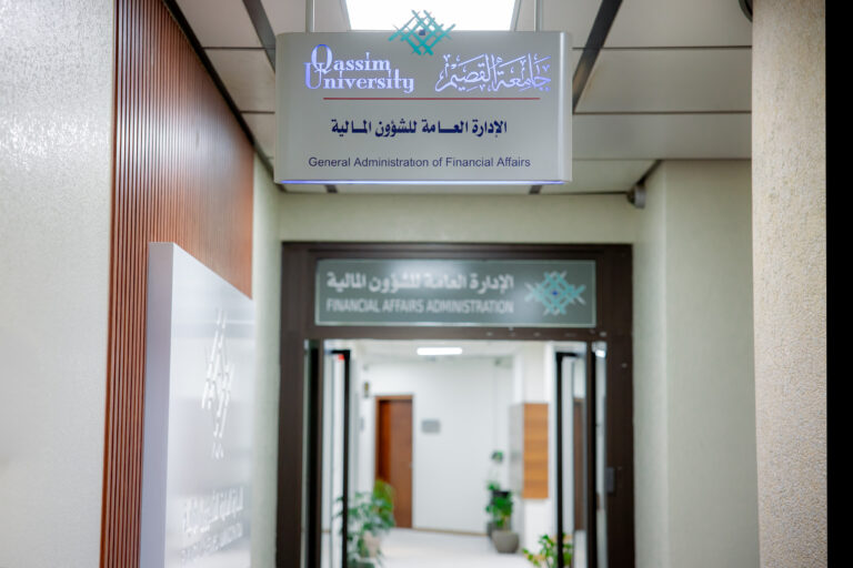 In one of the corridors of Qassim University, there is an overhead sign that reads "General Administration of Financial Affairs" in both Arabic and English, directing visitors to the financial management office. There are individuals dressed in traditional Saudi attire, such as a thobe with a shemagh or ghutra. The atmosphere reflects a formal and professional university environment.