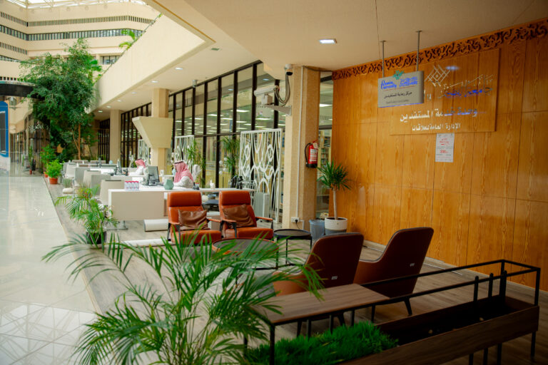 The image shows a modern interior office area decorated with green plants, Bengali chairs, and wood-paneled murals displaying an official Arabic text about public relations. A number of people wearing the Saudi thobe and shemagh or Saudi ghutra sit behind desks to provide customer service, and large windows allow natural sunlight to enter the space.