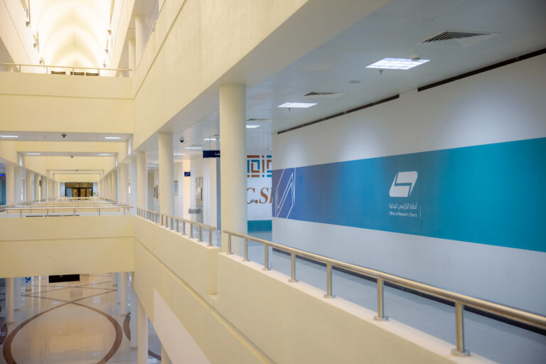 The modern and spacious corridor is characterized by its white walls, columns and elegant handrails, and the blue and pink colored signboards add a distinctive touch on the right wall, where the words "Research Chairs Secretariat" clearly stand out in both Arabic and English The space is well-lit and has a modern design that reflects the university environment of Al-Qassim University.
