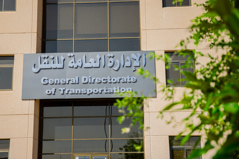 It is topped with a sign bearing the name of the General Directorate of Transportation in both Arabic and English, surrounded by green plants in the foreground.