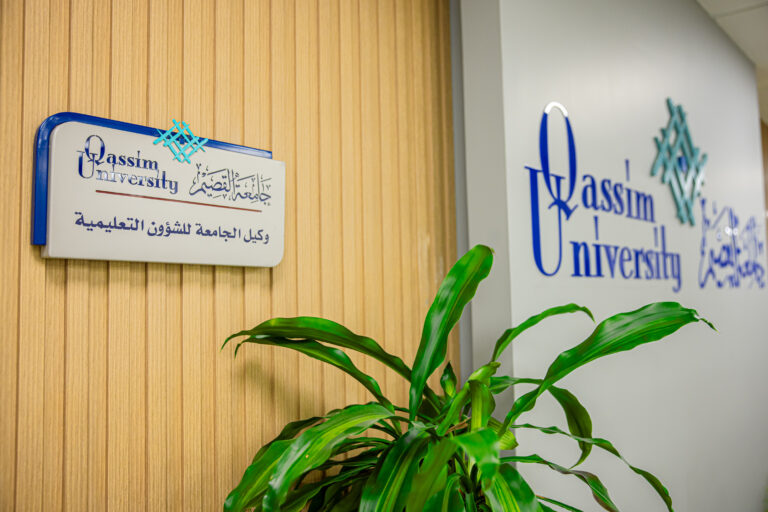 A plant placed in front of a wall bearing the logo of Qassim University and a pattern in Arabic, as well as a plaque indicating the university's Office of Educational Affairs.