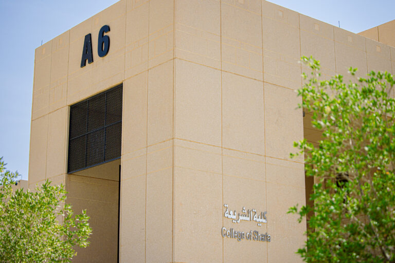 مبنى ذو لون بيج يحمل الرمز أ6 واسم كلية الشريعة باللغتين الإنجليزية والعربية، ويحيط به أشجار خضراء تحت سماء صافية زرقاء.