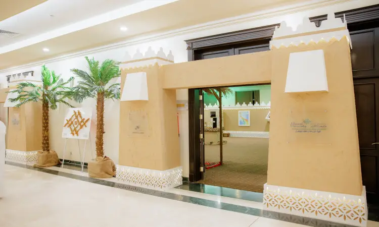 مدخل مبنى متميز لحداثة معمارية تقليدية عربية، يبرز من خلال جدرانه لامعة ونقوشه هندسية، تتوزع حوله نخيل صناعية عصر عصر التراث، ويزين المدخل لافتتان زجاجيتان يحملان نصاً عربياً بالقرب من باب لمعرض الجامعة في كرنفال بريدة للتمور. يظهر أمام المبنى عددٌ من الزوار مرتدين الشماغ السعودي والعقال والبشت أو الغترة السعودية، مما يدل على هوية المنطقة الرسمية ويرسِّخ قيم التراث التي تفتخر بها جامعة القصيم.