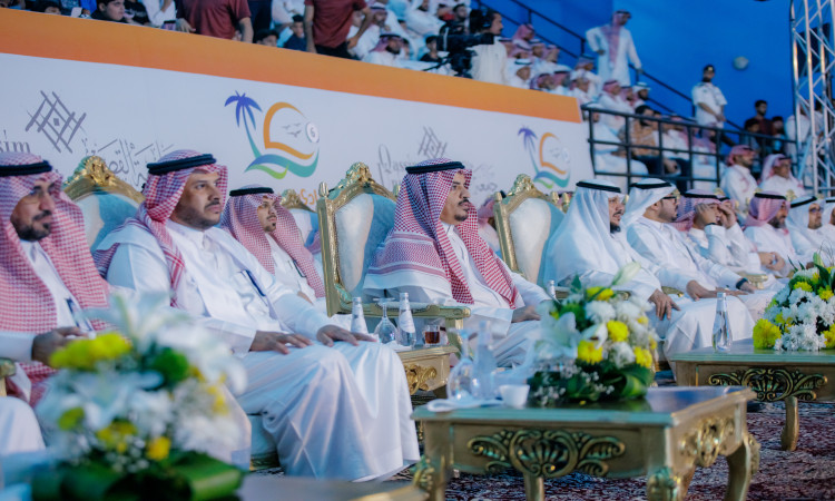 يجلسون مجموعة من الرجال مرتدين الغترة أو الشماغ والعقال، ومجتبين بالبشوت السعودية، يتقدمون رئيس الجامعة، على مقاعد البدلاء خلال ختام النادي الصيفي السادس في جامعة القصيم. وتزيّن الطاولات أمامهم بأكياس الزهور المتنوعة، بينما يظهر في حضورٌ كثيف يملأ قاعة الاحتفالات.