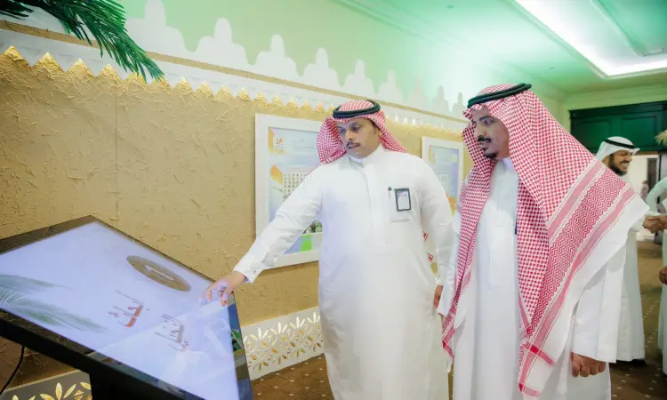 يتفاعل رجلان يرتديان الزي السعودي الرسمي الامتداد في الغترة أو الشماغ والعقال بالإضافة إلى البشت السعودي مع شاشة تفاعلية كبيرة في جناح جامعة القصيم خلال كرنفال بريدة للتمور، وجزء في الخلفية عدد من الحضور وبعض العناصر الزخرفية.