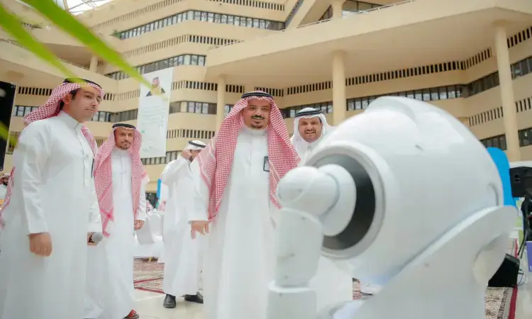يقود رئيس جامعة القصيم مجموعة من الرجال الحاضرين الغترة أو الشماغ والعقال والبشت السعودي، وهم يبتسمون ويتفاعلون مع روبوت بشري أبيض داخل مبنى حديث، في مشهد يجسد روح الرقمية خلال فعاليات يوم التوعية.