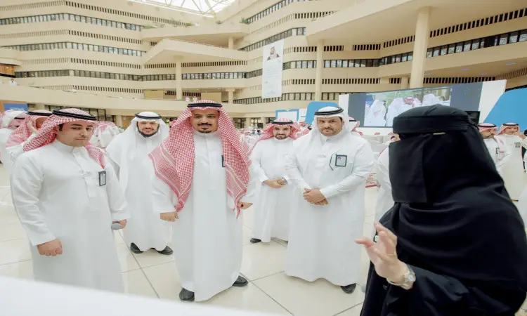 مجموعة من الرجال الذين يرتدون الغترة أو الشماغ مع العقال والبشت السعودي، بالإضافة إلى رئيس الجامعة، يستمتعون باهتمامهم بملابس أبلة ونقابًا أسود، وذلك في فعالية داخلية مخصصة لموضوع التلفاز الرقمي. وقد أدى هذا التنوع المعماري الجديد إلى ظهور ظاهرة كبيرة في الخلفية.