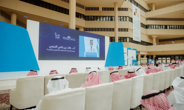 قاعة مؤتمرات تتسع لصفوف الكراسي البيضاء، يجلس عليها مجموعة من الرجال مرتدين الغترة أو الشماغ والقالب، ويزين الاشتراك البشت الرسمي السعودي، ويدركون فعاليات "يوم التوعية" حول المؤتمر الرقمي. قبل حضوره شاشة كبيرة تحمل صورة معالي رئيس جامعة القصيم ونص عربي ذي طابع رسمي ضمن هذا الحدث الجامعي الهام.
