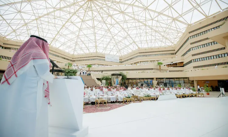 رجل يلقيٌ يرتدي أزياء سعودية قديمة متمثلة في الغترة والعقال والبشت السعودي كلمةً من منصة الخطابة، وذلك في بهو واسع وحديث يتميز بسقفٍ زجاجي مقبب، خلال فعاليات برنامج طموح. يجلس في صفوف الطلاب وأعضاء هيئة التدريس من جامعة القصيم منصتين باهتمامهم إلى المكان الصحيح.