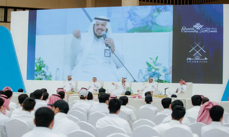 يجلس خمسة رجال تحت الغترة أو الشماغ والعقال والبشت السعودي على منصة في جامعة القصيم خلال فعالية برنامج طموح، ويتألقون أمام جمهور الطلاب ويعينون الجالسين على كراسٍ بيضاء. وقم بعرض خلفهم شاشة كبيرة للبحث والشعارات الصحيحة.