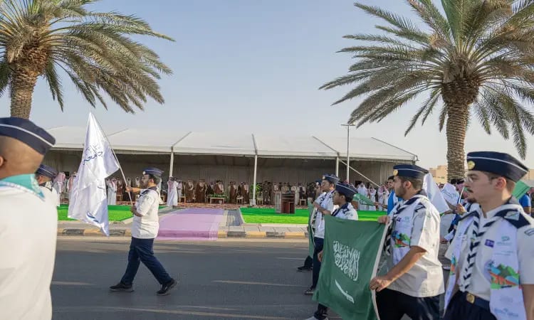 مجموعة من الكشافة بزيهم الرسمي، مرتدين الشماغ السعودي والعقال والبشت السعودي، يسيرون في موكب مهيب في الهواء الطلق ويحملون علم المملكة العربية السعودية ولافتات احتفاءً باليوم الوطني 93. وتحيط بهم النخيل وخيمة كبيرة ونصبت بجوار الجامعة. ويشاهد عدد من الحضور يتقدمهم سمو أمير المنطقة من داخل الخيمة بالقرب من جامعة القصيم.