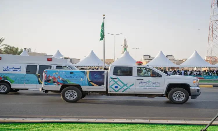 تمر سيارة نقل مزينة وسيارة إسعاف أمام خيام تحكم بشكل كبير خلال فعالية عامة لليوم الوطني 93 للمملكة العربية السعودية، حيث يرفرف العلم السعودي عاليًا إلى النهاية. يشارك ثلاثة طلاب من جامعة القصيم في الاحتفال، ويرتدون الغترة أو الشماغ والعقال والبشت السعودي، مجسدين مظاهر الفخر والانتماء للوطن في هذه الأهداف الوطنية العظيمة.