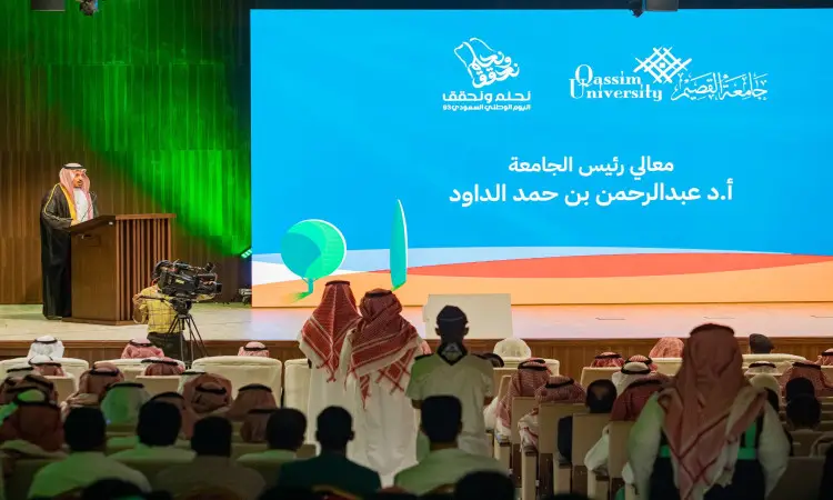 He takes the podium in front of the audience at the event, with a large screen behind him bearing the Qassim University logo and a text in Arabic about National Day. In the hall, a number of attendees are wearing shemagh or ghutra and headband, in addition to the Saudi bisht, as a sign of national identity and the official traditions of the occasion.