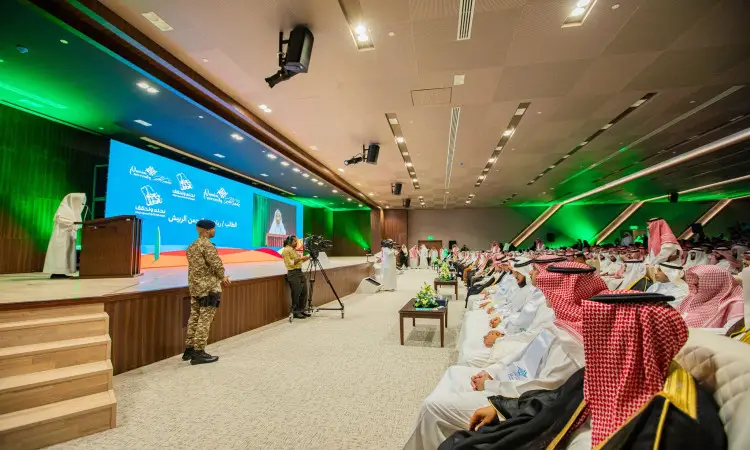 The conference hall is lined with men wearing the Saudi Ghutra or Shemagh with Eqbal, a number of them wearing the official Saudi bisht, all standing on a podium with a blue screen. The emir of the region delivers his speech from the podium, surrounded by the official leaders. The green stadium adds a formal touch to the National Day celebration at Qassim State University.